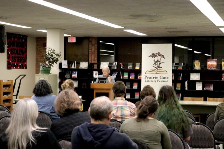 A speaker at a podium addresses a small audience in a library during the Prairie Gate Literary Festival. Shelves of books and a festival banner are visible in the background, while attendees sit attentively in rows of chairs. A large tapestry with the words 'No More Stolen Sisters' hangs on a wall to the left.